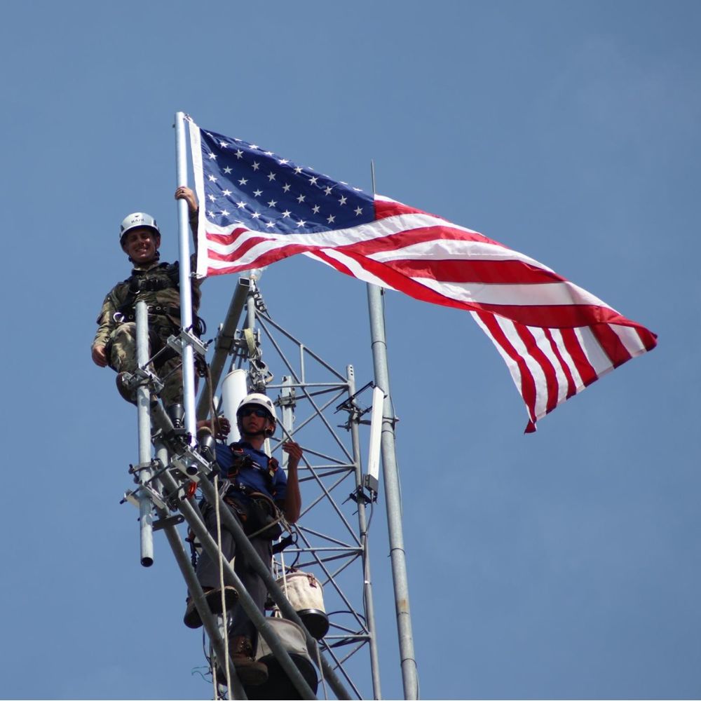 MetaLINK technicians standing on a telecommunications tower proudly flying the American flag against a clear blue sky