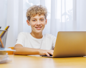 Young boy with curly blond hair smiling while doing homework on a laptop at a desk during the school year