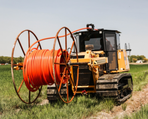 A yellow tracked fiber optic cable laying machine with a large spool of bright orange cable sits in a grassy rural field, ready for underground internet installation.