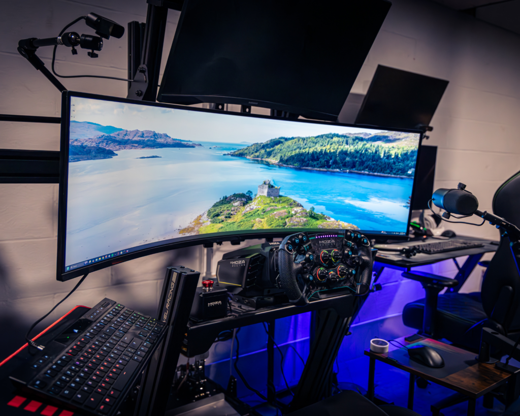 Modern gaming desk setup with ultrawide curved monitor, Moza Racing sim wheel, RGB mechanical keyboard, headset mount, and blue under-desk LED lighting in a home office space.