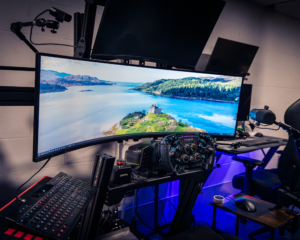 Modern gaming desk setup with ultrawide curved monitor, Moza Racing sim wheel, RGB mechanical keyboard, headset mount, and blue under-desk LED lighting in a home office space.