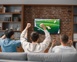 Three friends sit on a couch watching soccer on TV mounted on a brick wall, cheering with raised arms as their team scores a goal.