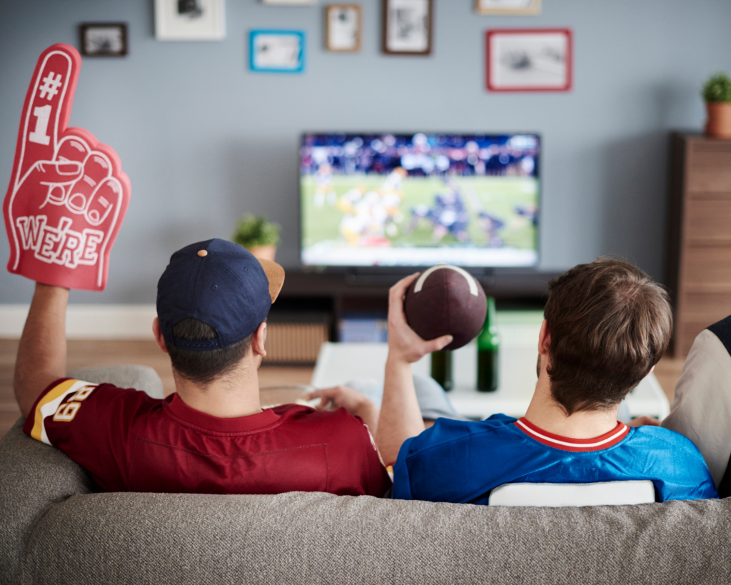 Two friends in football jerseys sit on a couch watching a game on TV, one holding a foam finger and the other holding a football.